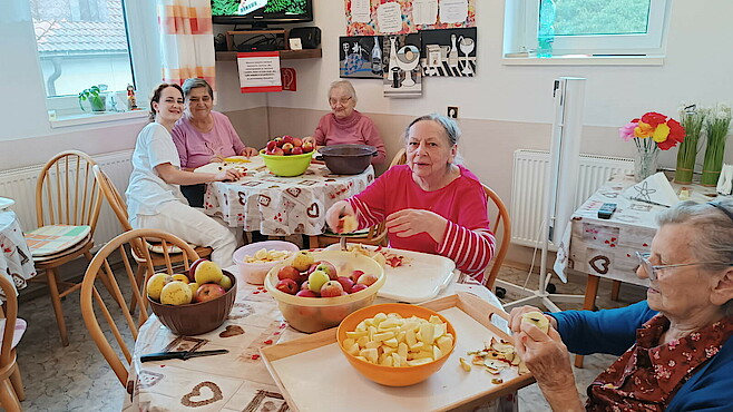 Vier Seniorinnen und eine Pflegerin sitzen an zwei Tischen, schälen Äpfel und schauen in die Kamera.