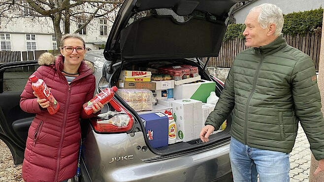 Eine Frau und ein Mann stehen vor einem mit Lebensmitteln beladenen Auto. Der Kofferraum ist offen und man kann die Lebensmittel sehen.
