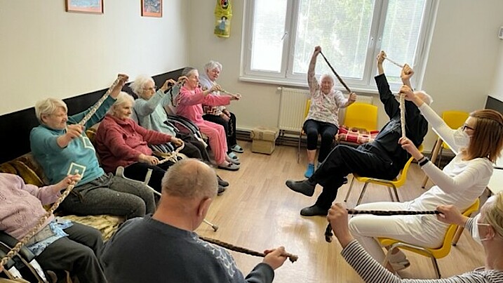 Senorinnen und Senioren sowie eine Pflegerin sitzen auf Stühlen oder einem Sofa in einem Raum und machen Physiotherapie.