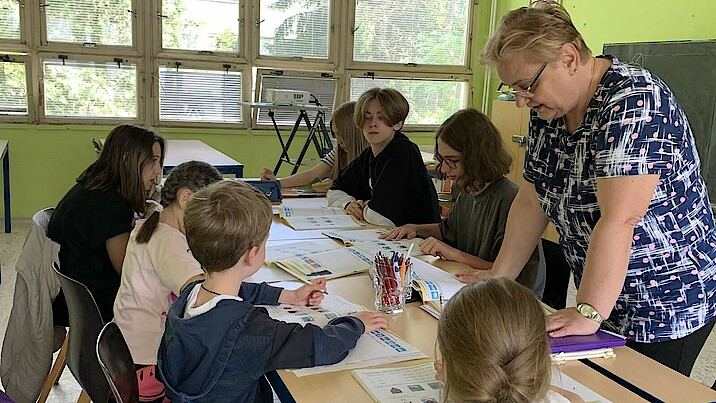 Sechs Kinder und eine Lehrerin stehen und sitzen an einem Tisch und lernen.